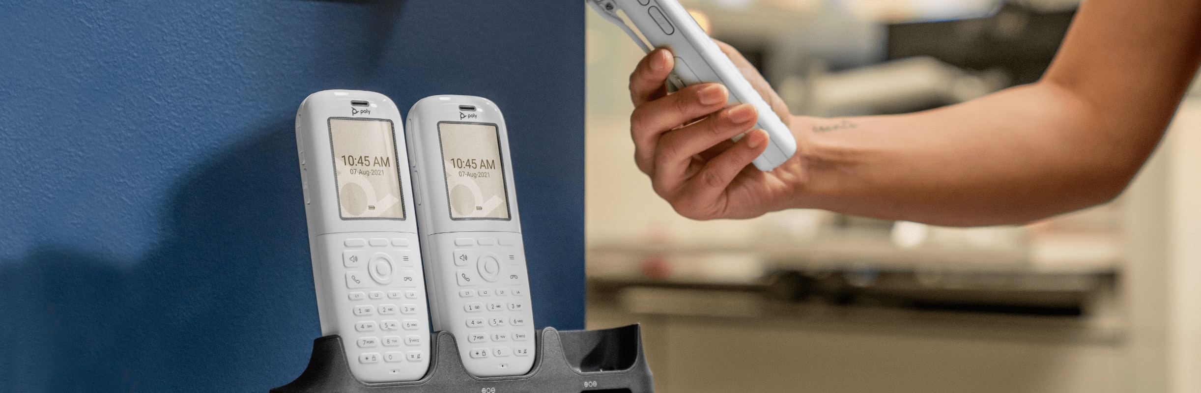 Person in office, holding a Poly Rove 40 wireless IP phone handset while two Poly Rove 40 handsets remain in base station beneath wall-mounted DECT repeater