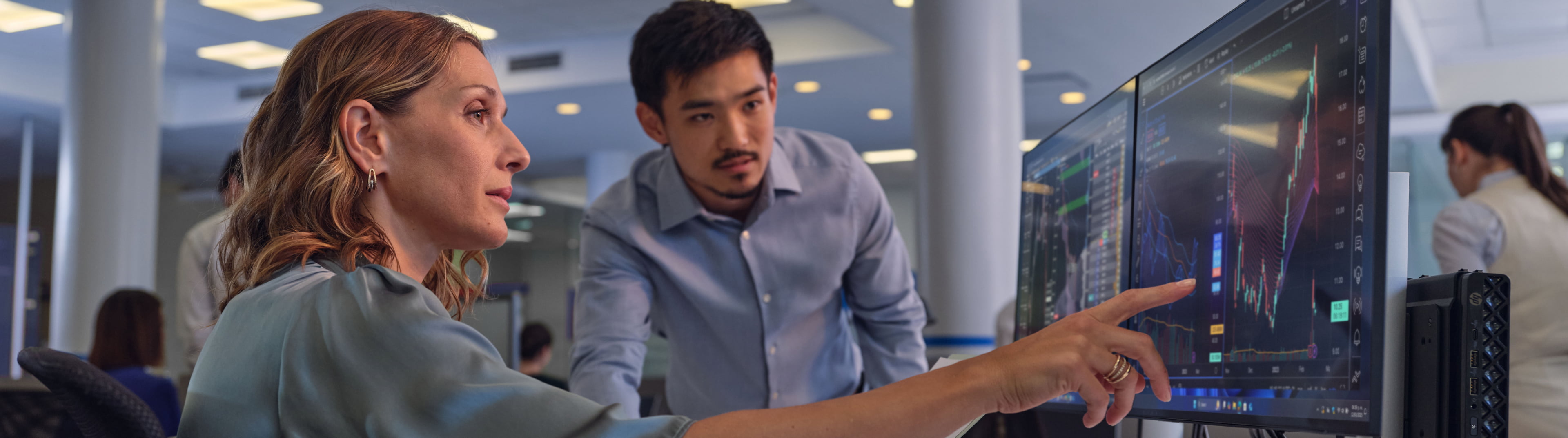 female IT professional pointing at data on dual monitors teaching young male colleague